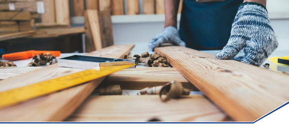 A person wearing safety gloves measures wooden planks on a workbench. Nearby are a measuring tape, pencil, and wood shavings, conveying craftsmanship.