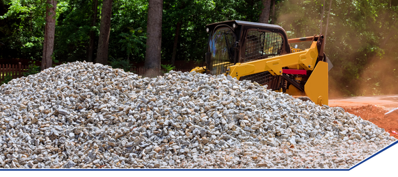 Bulldozer moving a large pile of gray gravel in a forested area. Sunlight filters through the trees, creating a busy, industrious atmosphere.